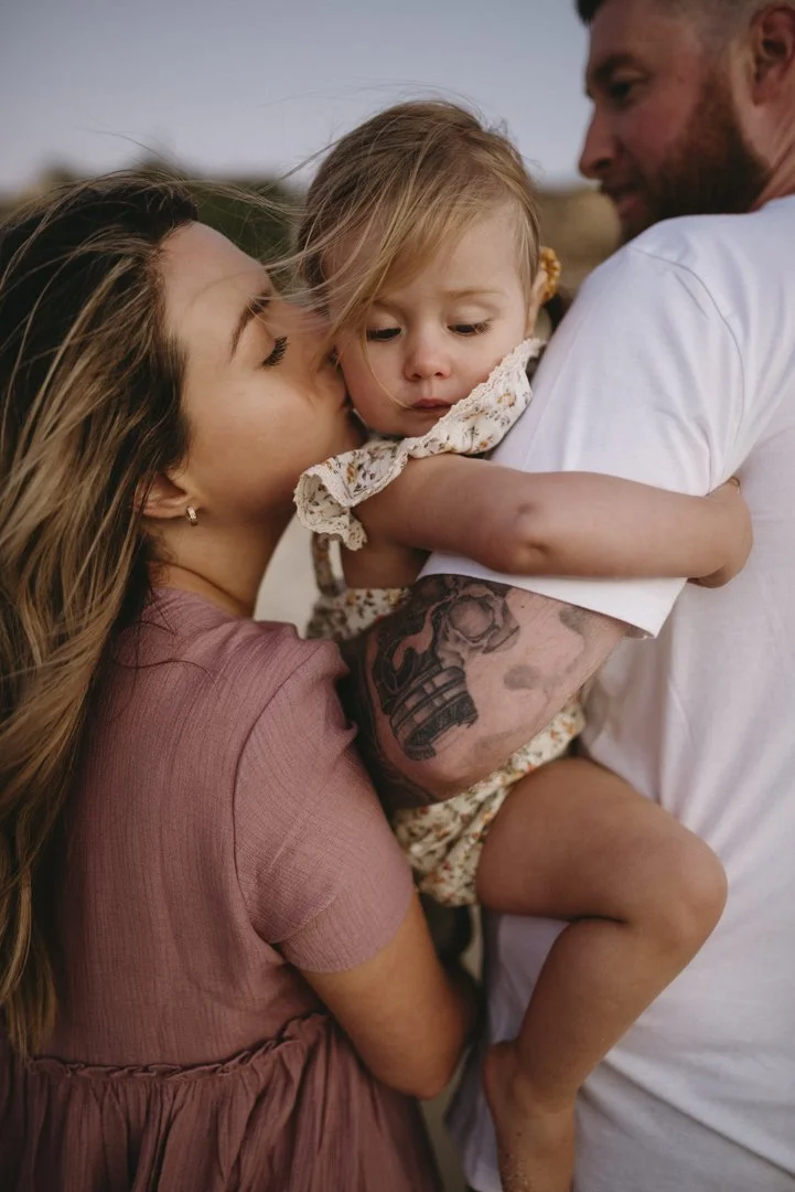 A woman kissing a child's cheek, who is being held by a man with a tattoo on his arm. The scene is outdoors.
