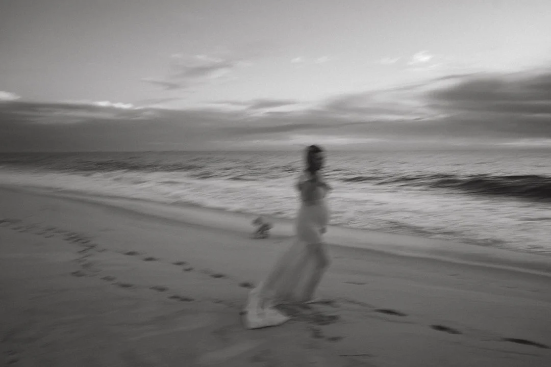 Blurred black and white image of a person walking on a beach with ocean waves and cloudy sky in the background.