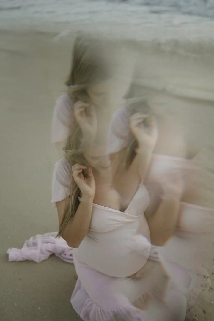 Pregnant woman in pink dress kneeling on sandy beach with a blurred effect.