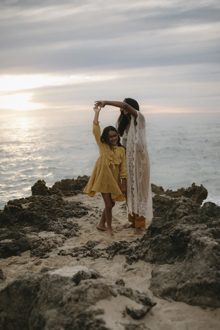 Two people on a rocky beach at sunset, one with raised arm holding the other's hand, both smiling and dressed in light, flowing garments.