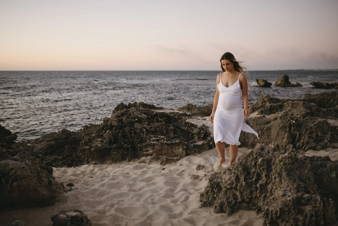 Woman in white dress walking on a rocky beach at sunset.