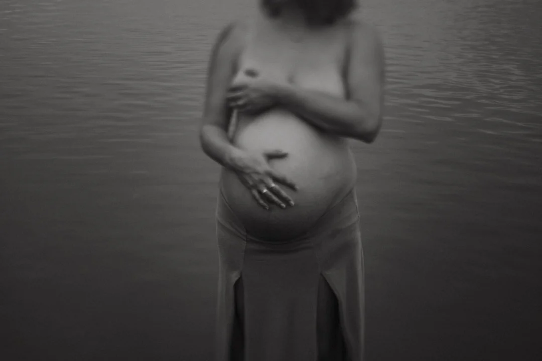 Black and white photo of a pregnant woman standing in water, covering her chest and holding her belly.