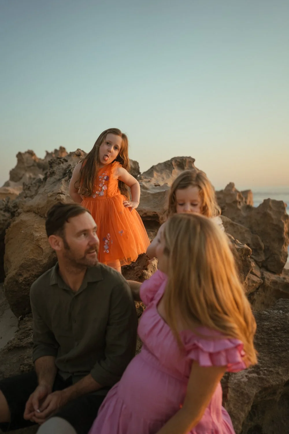 Family with two children on rocks at sunset; girl in orange dress sticks out tongue.