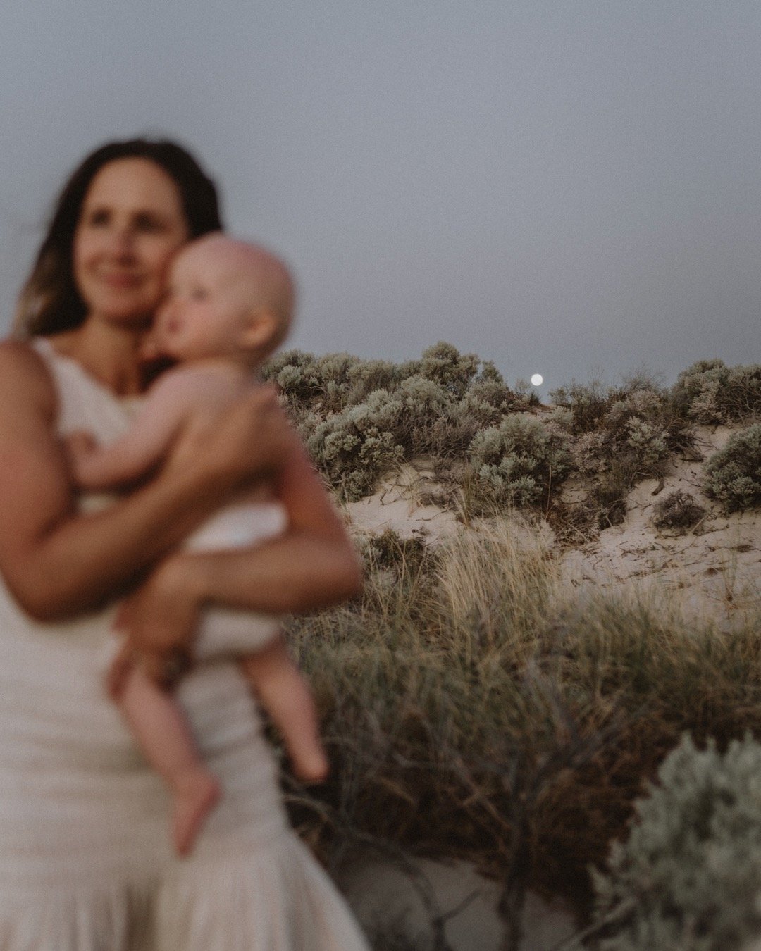 Blurred photo of a woman holding a baby with sand dunes and vegetation in the background, under a clear sky.
