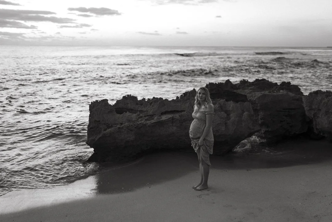 Pregnant woman standing on a beach near large rocks, with ocean waves in the background, captured in black and white.