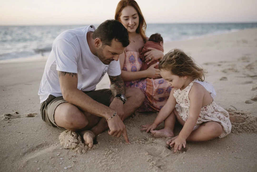 Family with two children sitting on a sandy beach, with a man pointing in the sand, a woman holding a baby, and a girl playing in the sand.