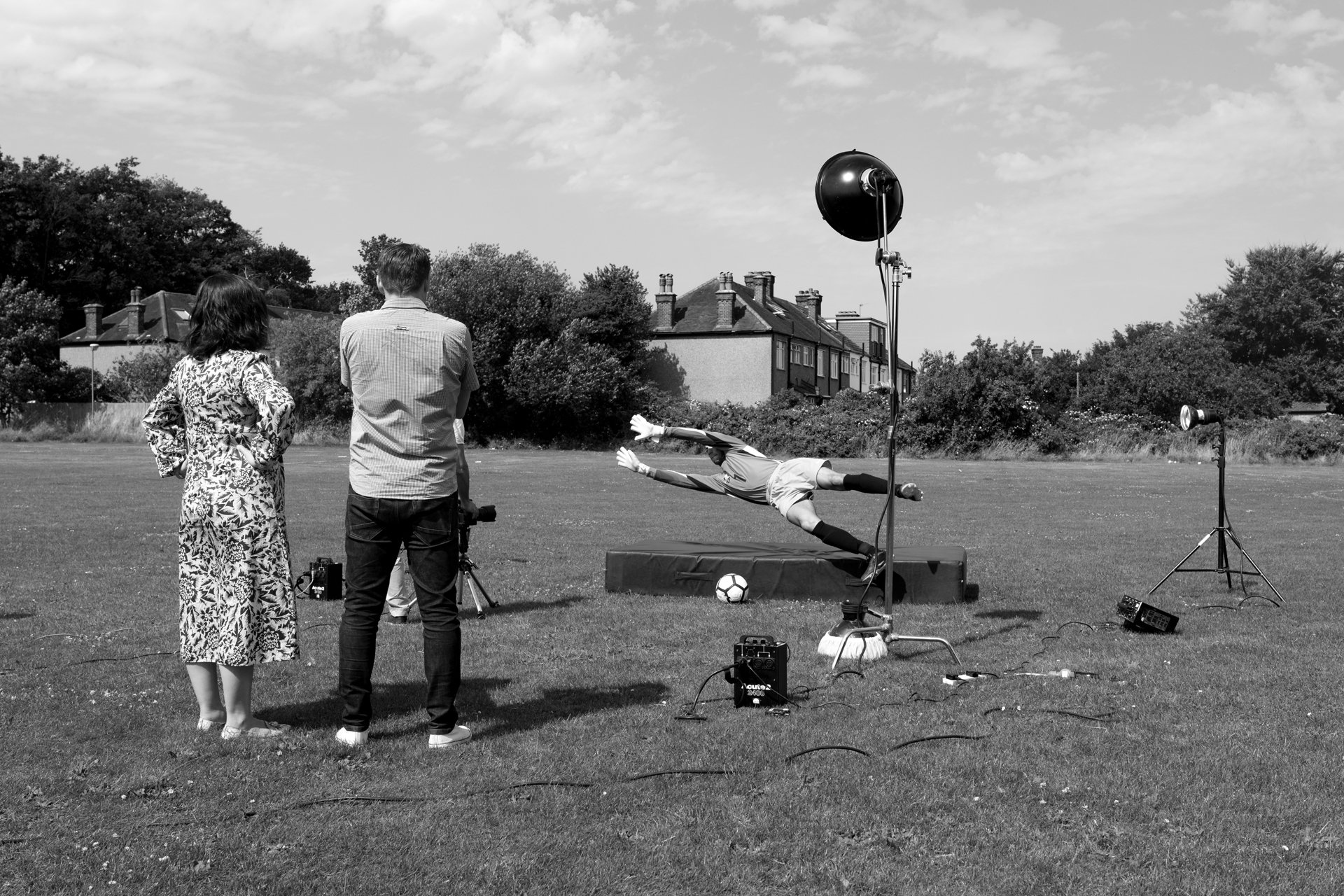 A black and white photo of an outdoor photoshoot with a soccer goalkeeper diving to save a ball. Two people, a woman in a floral dress and a man in a checked shirt, are standing observing. Studio lighting equipment and other setup tools are visible on the grass field, with residential houses and trees in the background.