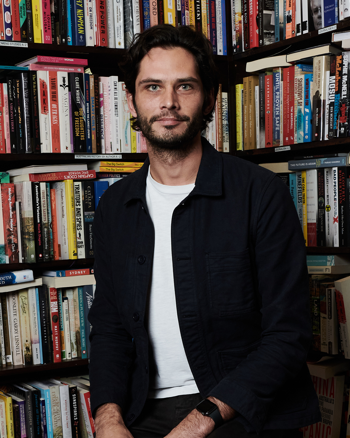 A man with dark hair and a beard sitting in front of a bookshelf filled with various books, wearing a black jacket and a white t-shirt.
