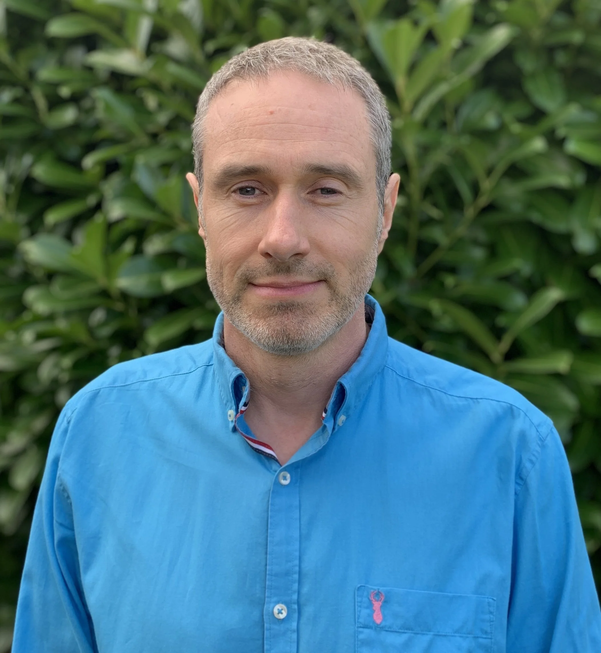 A man in his late forties with short grey hair, wearing a blue button-up shirt, standing outdoors in front of green foliage.