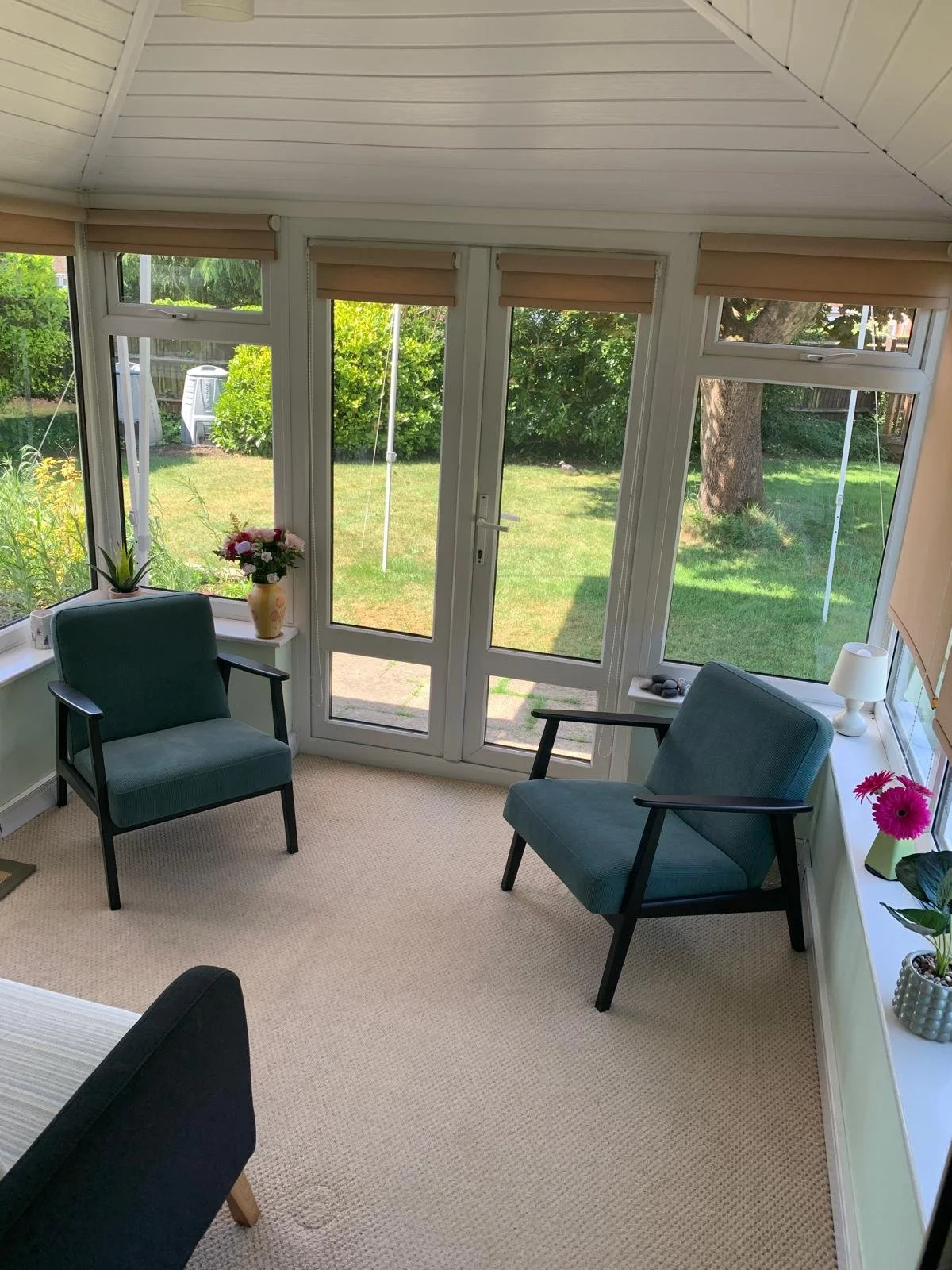 A counselling room with two teal armchairs, potted plants, vases with pink and white flowers, a small white table lamp, and windows with beige blinds overlooking a green garden.