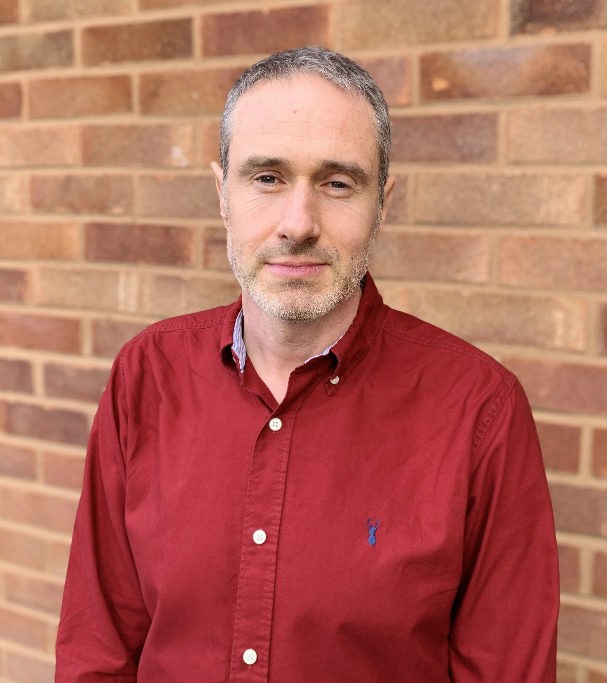 A man in his late forties with short grey hair, wearing a red button-up shirt, standing in front of a brick wall.