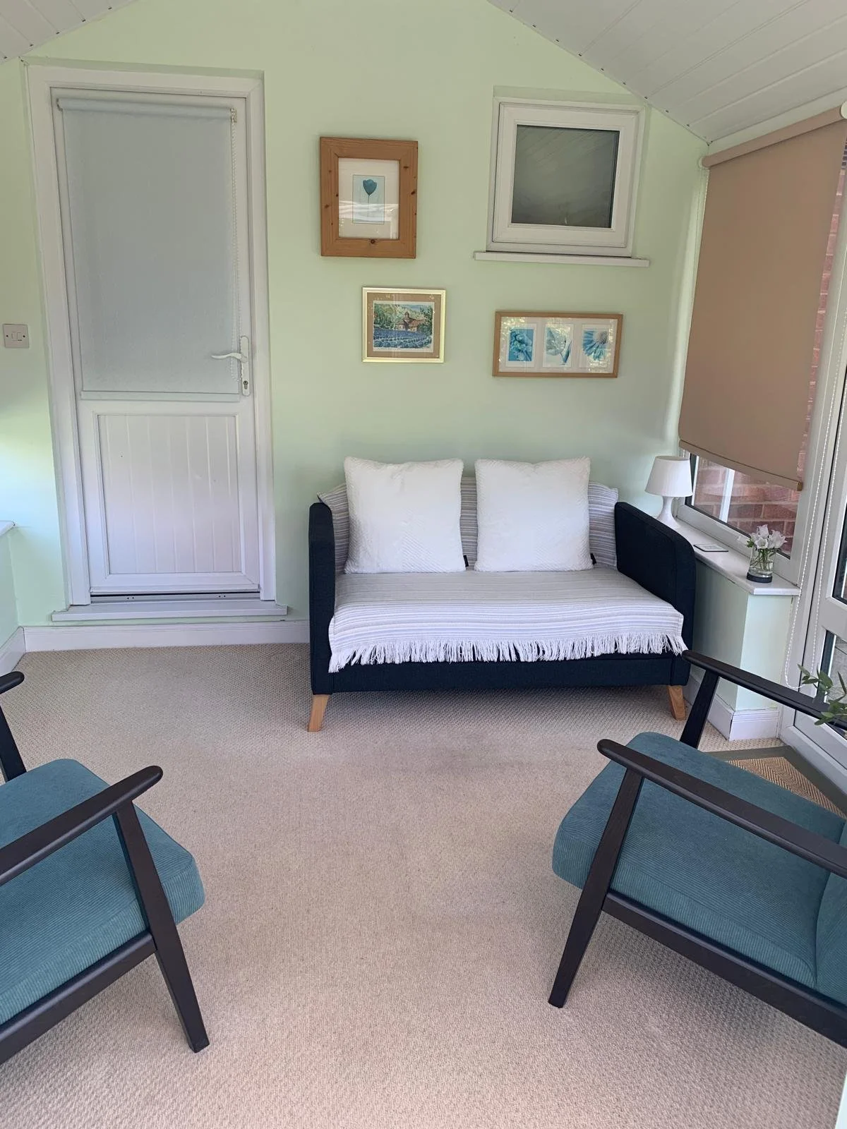 Counselling room with a small black sofa with white cushions, two teal armchairs, a lamp and a small plant, framed artwork on light green walls, a window with beige blind, and a glass door.
