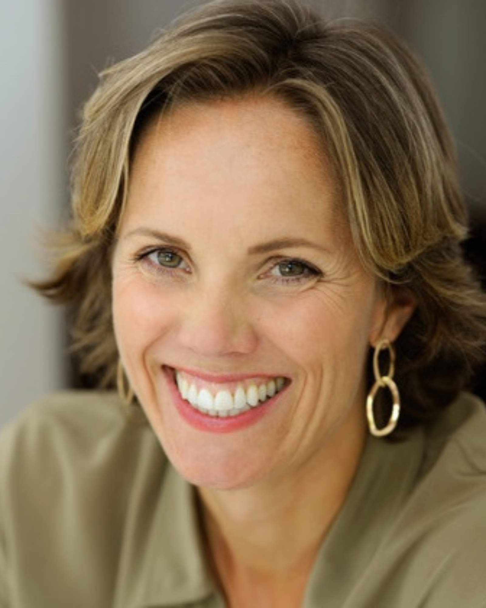 Close-up of a woman with short, light brown hair, wearing gold earrings and a beige top, smiling brightly.