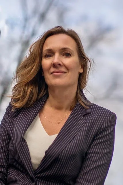 A woman with shoulder-length brown hair wearing a pinstripe blazer and a white shirt, standing outdoors against a cloudy sky.