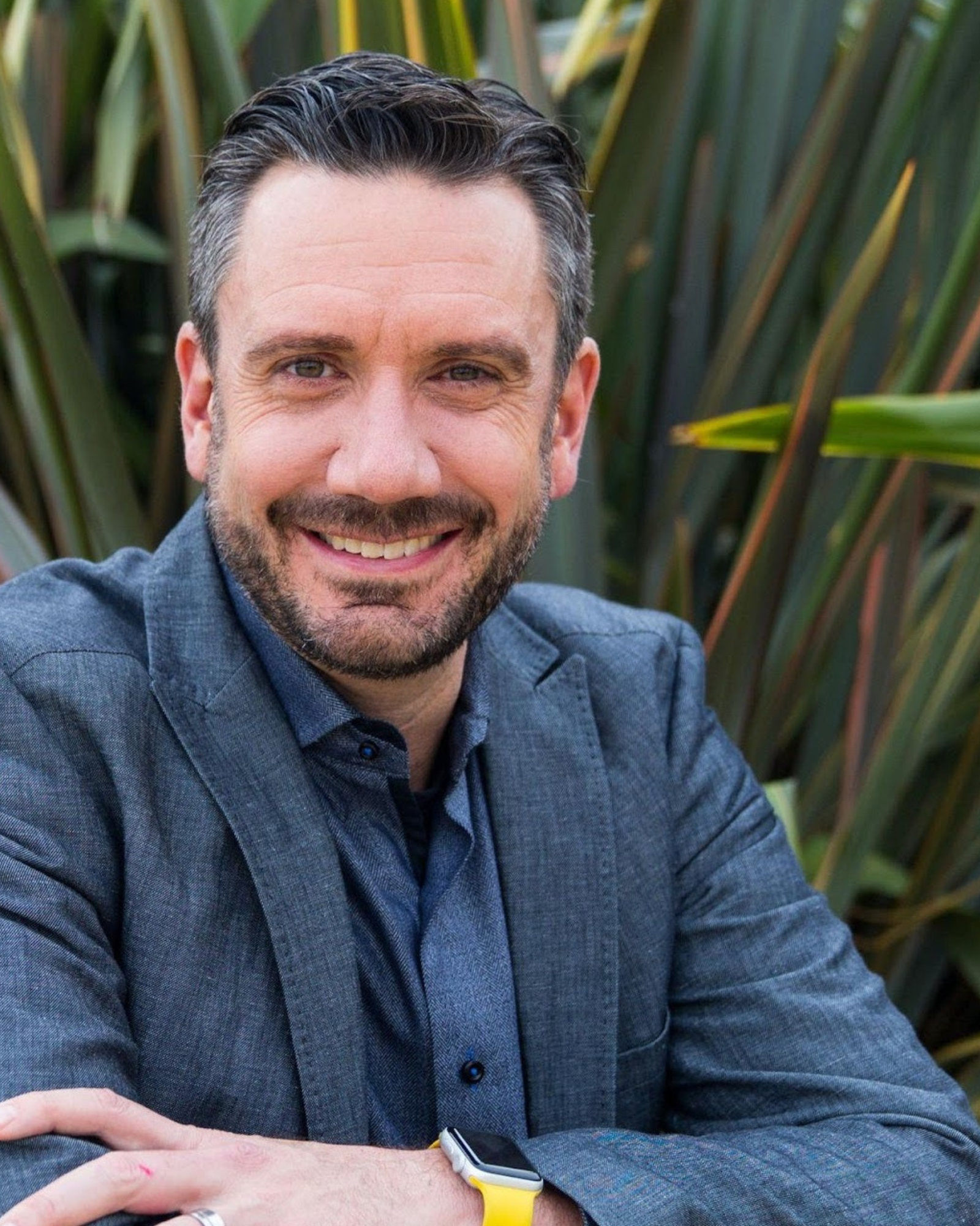 A smiling man with dark hair and a beard wearing a gray blazer and blue shirt, sitting outdoors with green plants in the background.