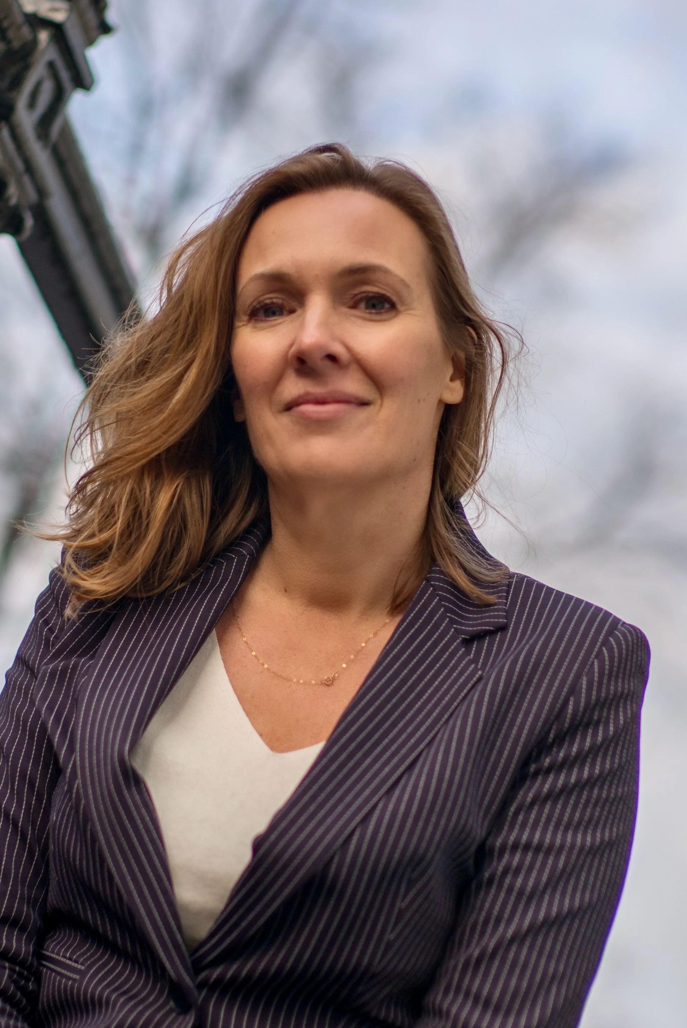 A woman with shoulder-length brown hair wearing a pinstripe blazer and a white shirt, standing outdoors against a cloudy sky.