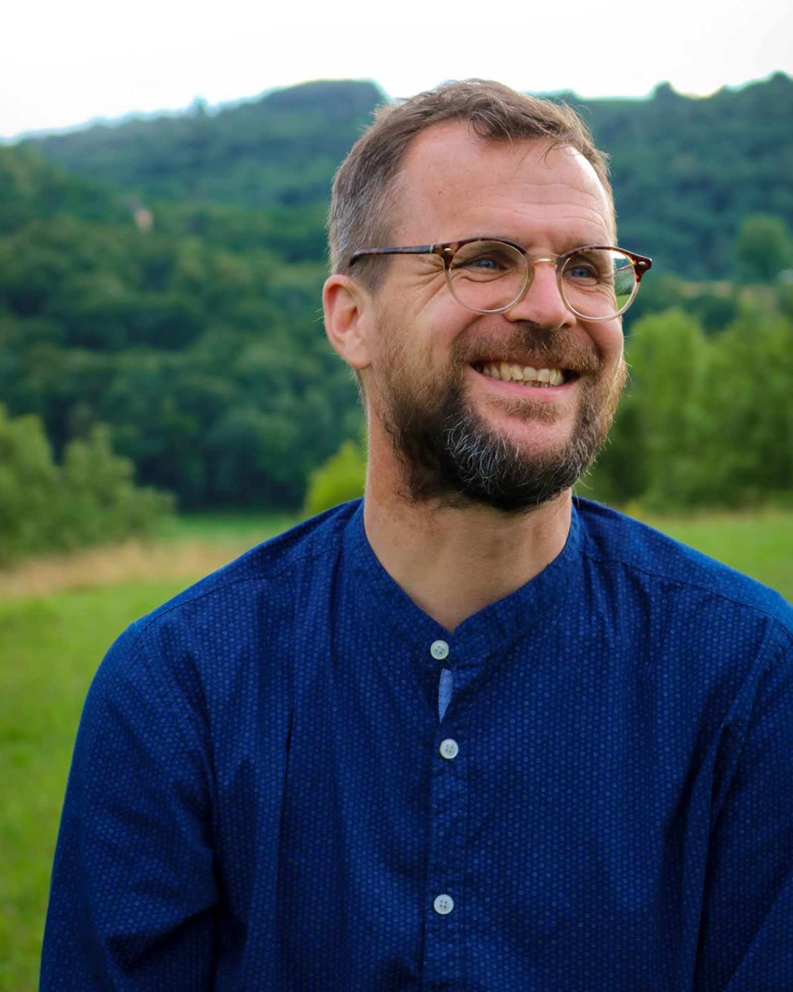Smiling man with glasses and beard outdoors in a green field, hills in the background.
