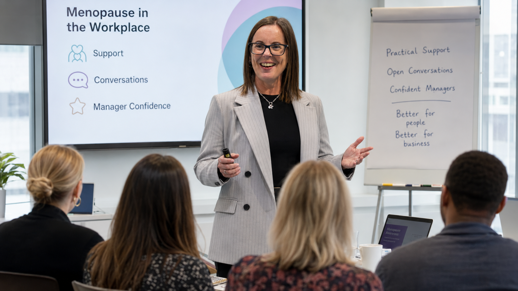 A woman in a gray blazer giving a presentation about menopause in the workplace to an audience in a modern office conference room.