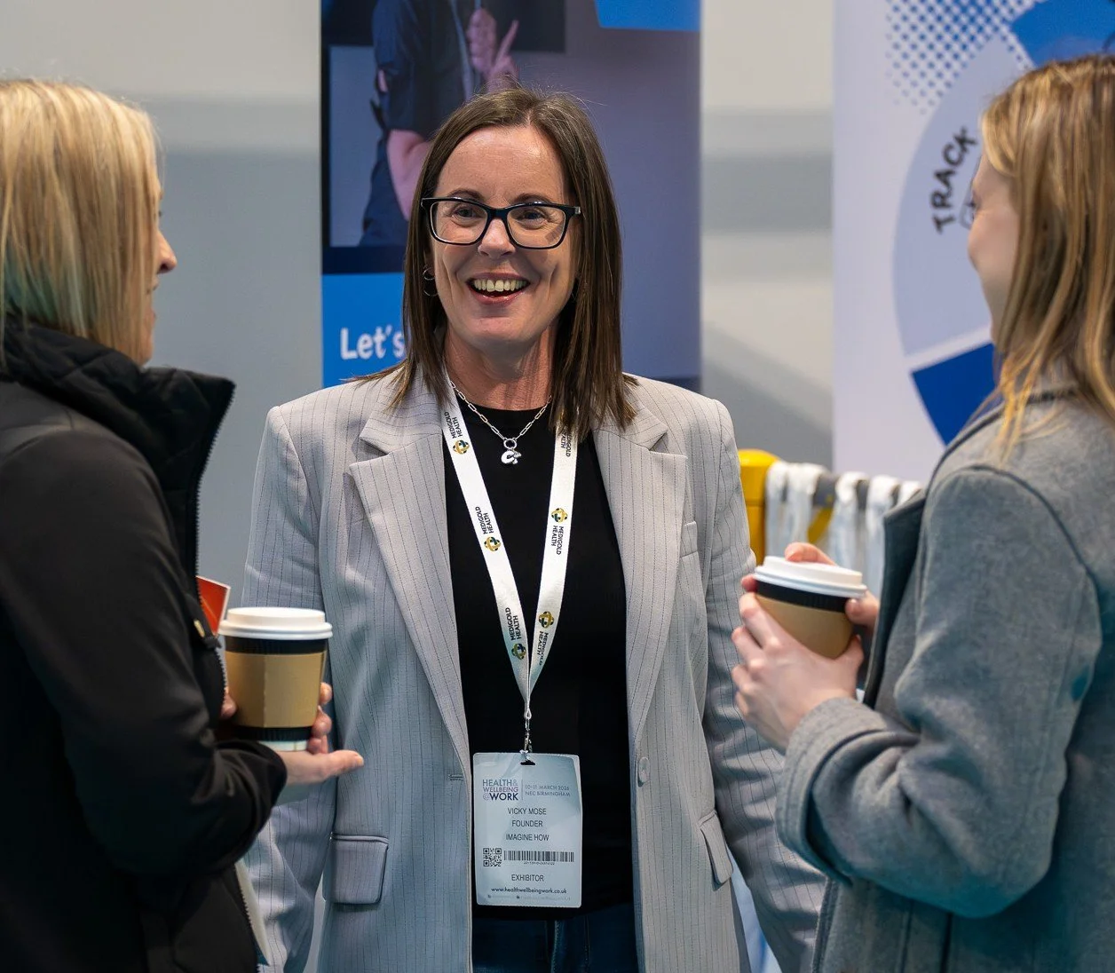 Three women talking at a conference, holding coffee cups, with a large badge and a display screen behind them.