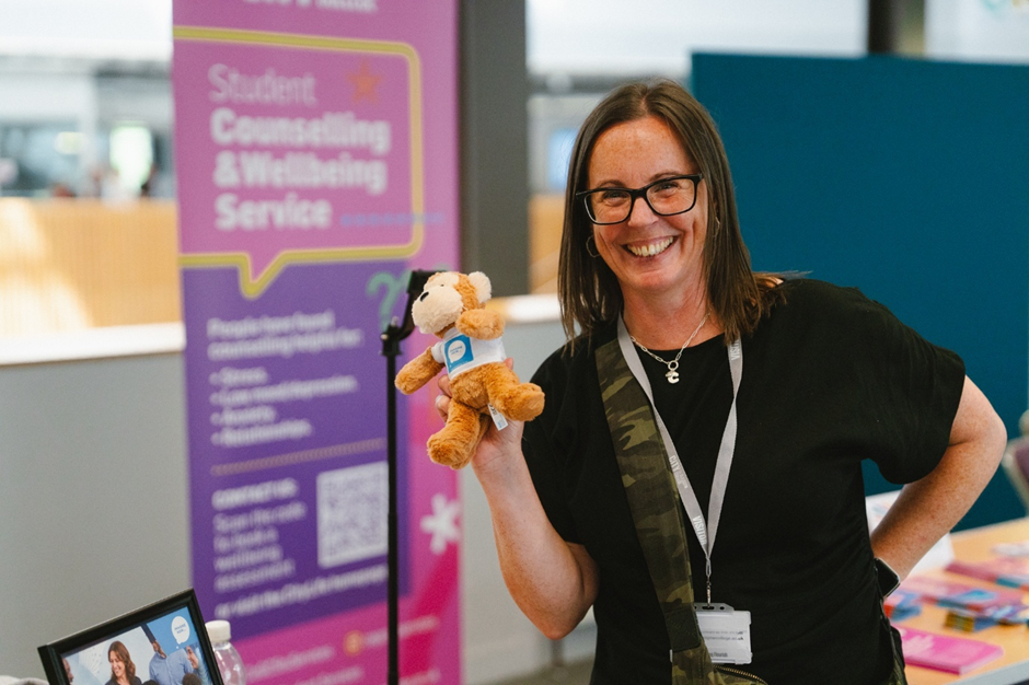 A woman with glasses smiling and holding a plush toy on a conference or event floor, with a purple banner and informational materials in the background.