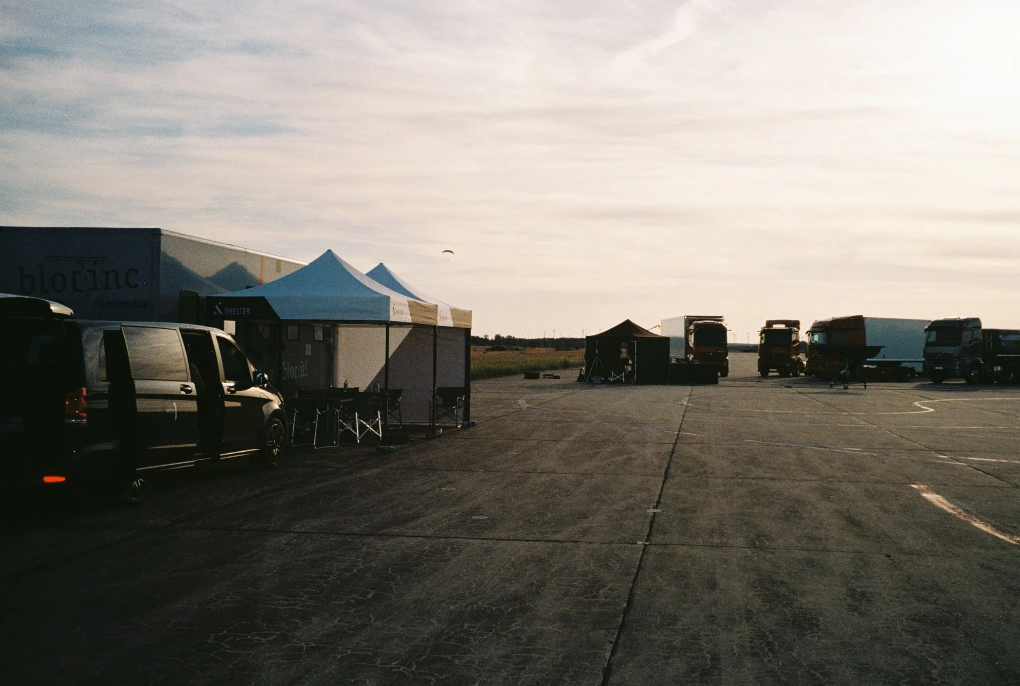 An outdoor scene at an airstrip with parked trucks, temporary tents, and a parked black van, under a partly cloudy sky with a small aircraft visible in the distance.
