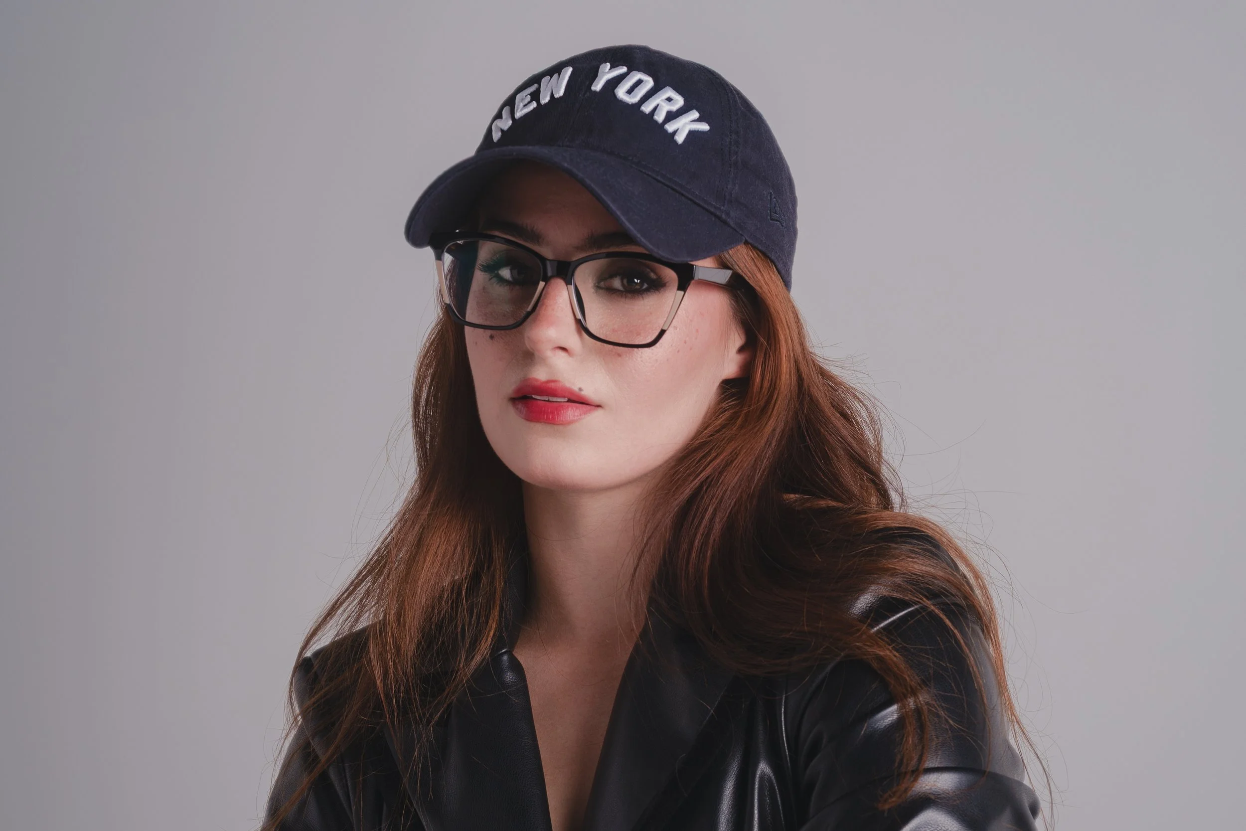Close-up lifestyle and apparel portrait of a model wearing a black leather jacket and a dark New York baseball cap. The image showcases fashion accessories, styling, and a bold, contemporary aesthetic.