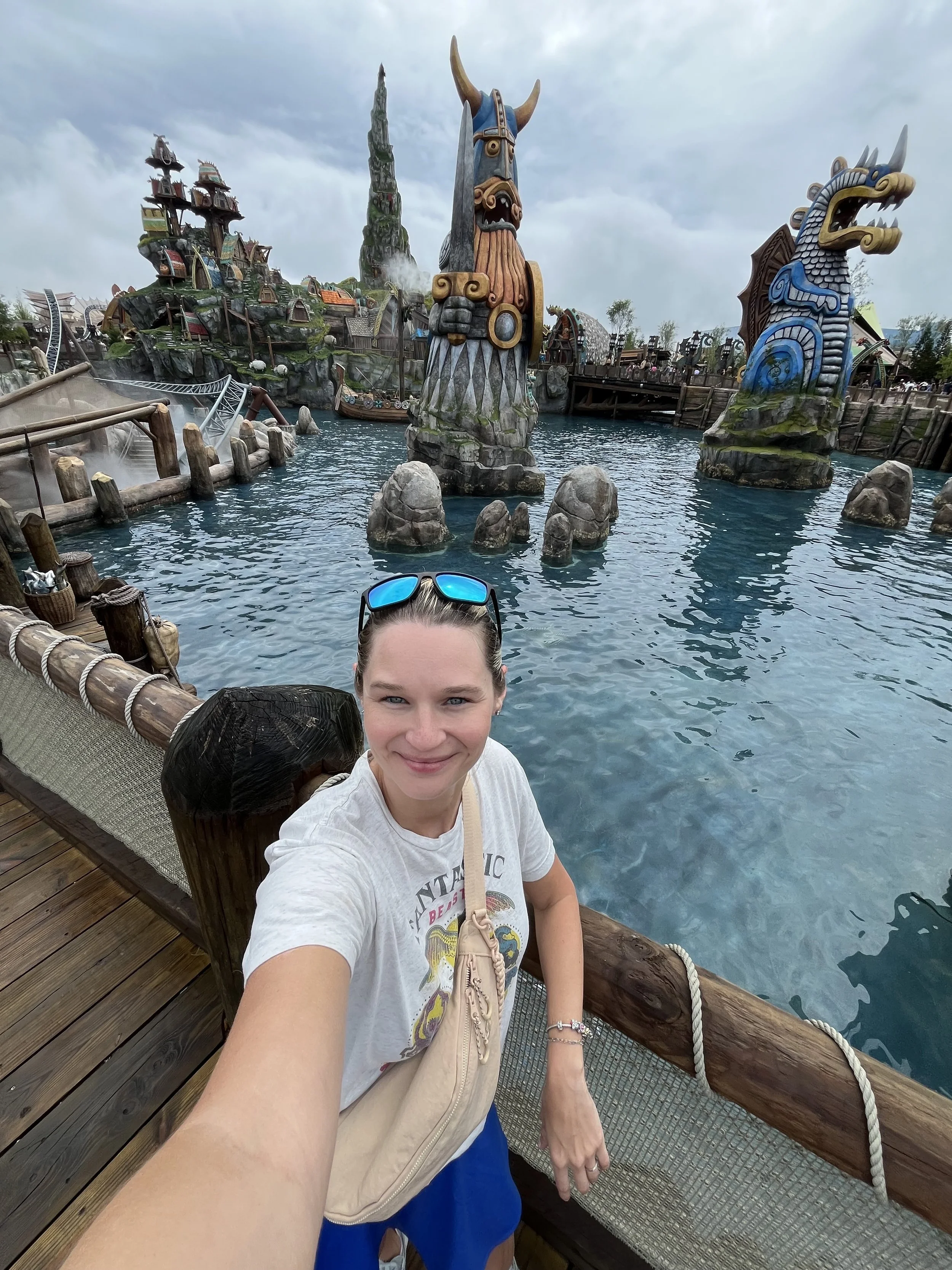 A young woman takes a selfie at a theme park, with Viking-themed statues and a roller coaster in the background.