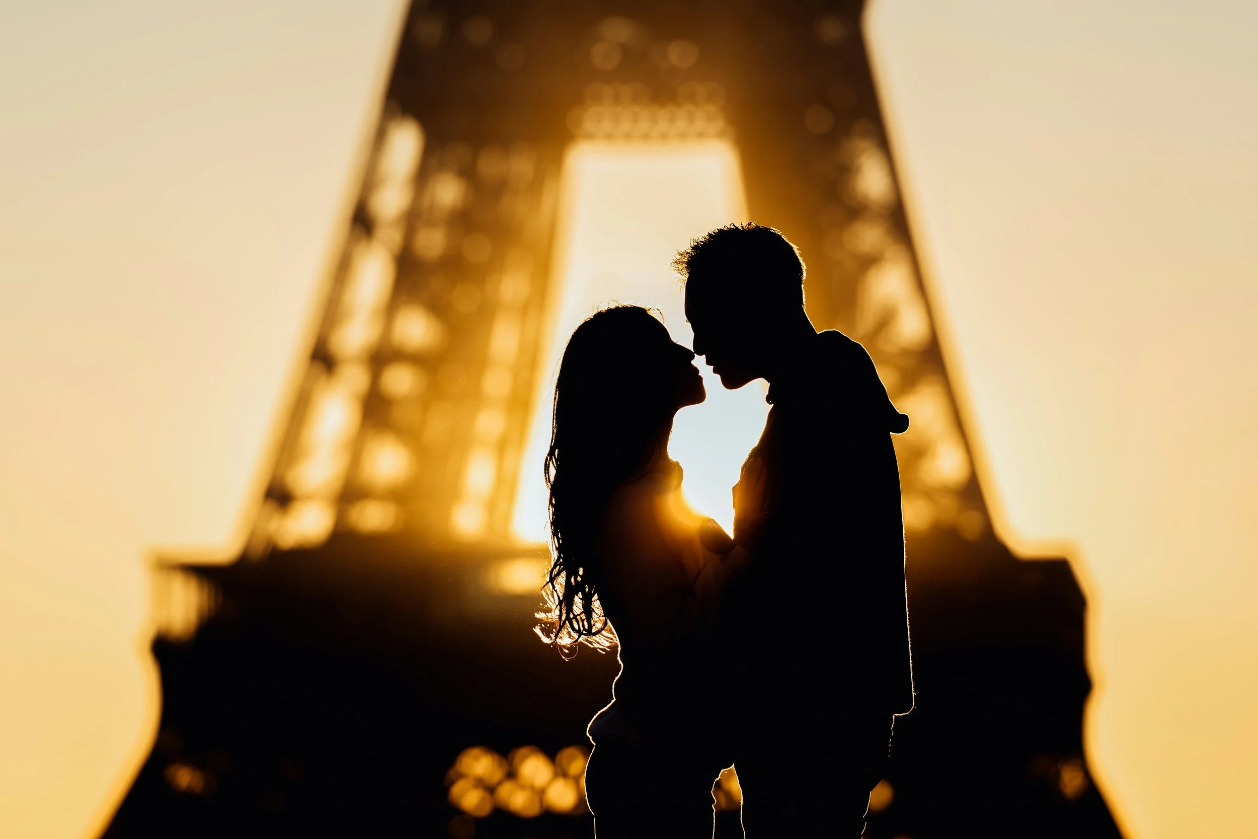 Silhouettes of a man and woman facing each other with their foreheads touching under the Eiffel Tower during sunset.
