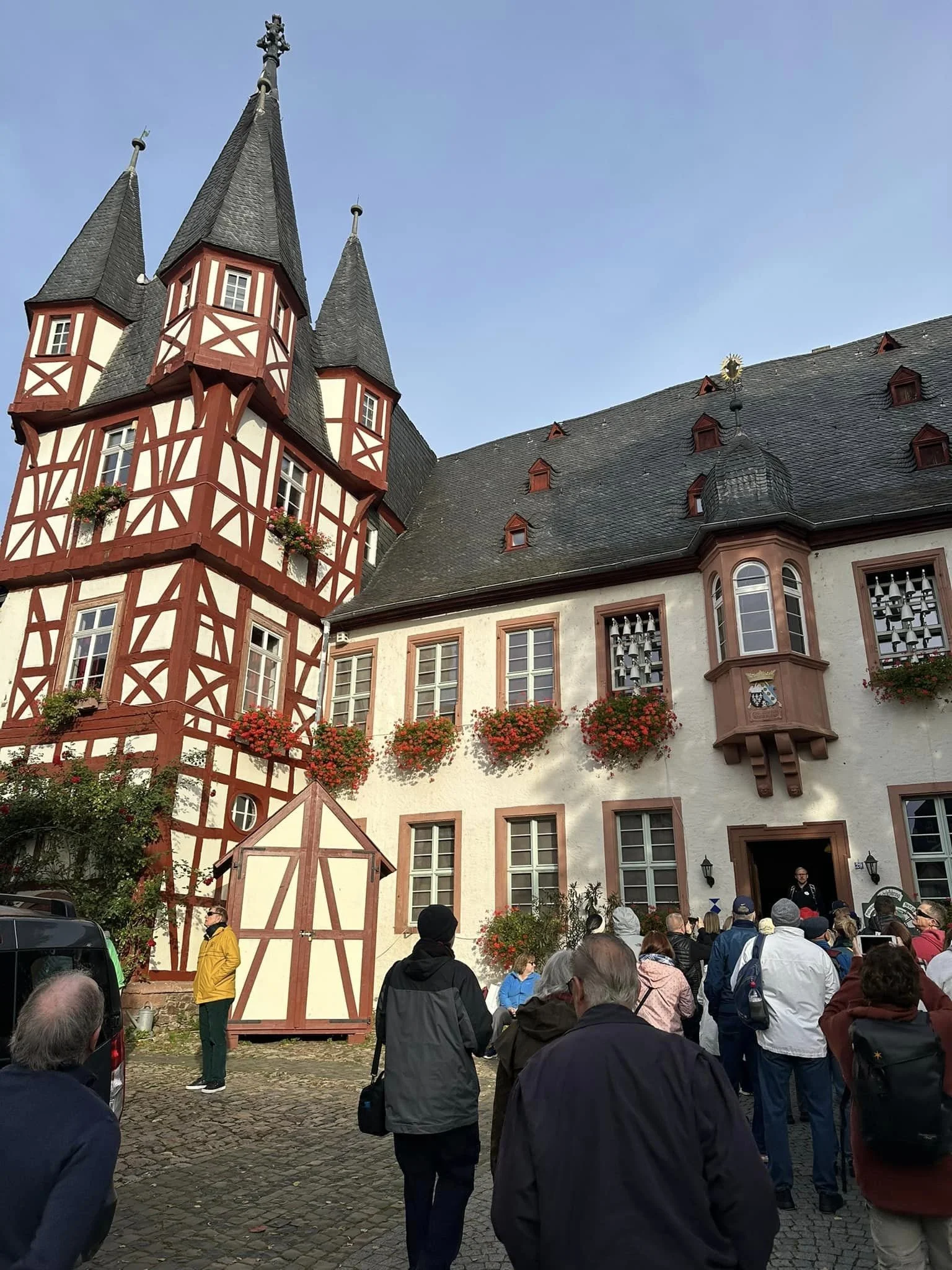 A crowd of people gathered in front of a historic European building with a steep roof, tower, and decorative wooden accents, during daytime.