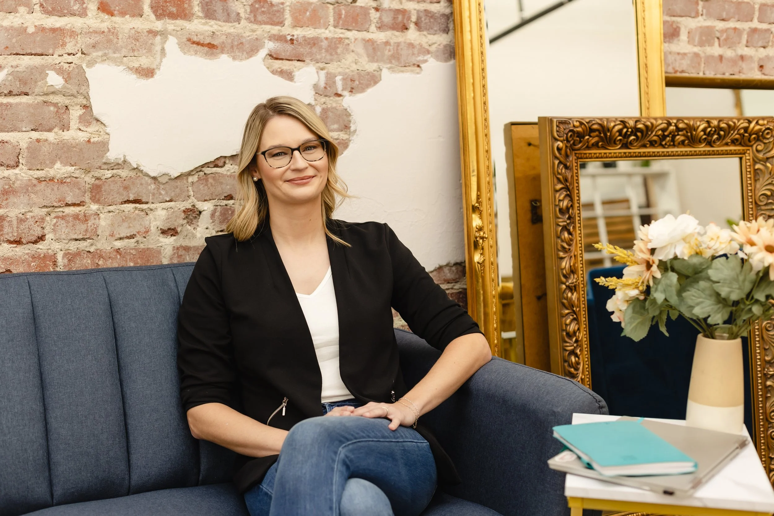 Woman with blonde hair and glasses sitting on a blue couch in front of a brick wall, with gold-framed mirrors and a table with a flower arrangement and notebooks nearby.