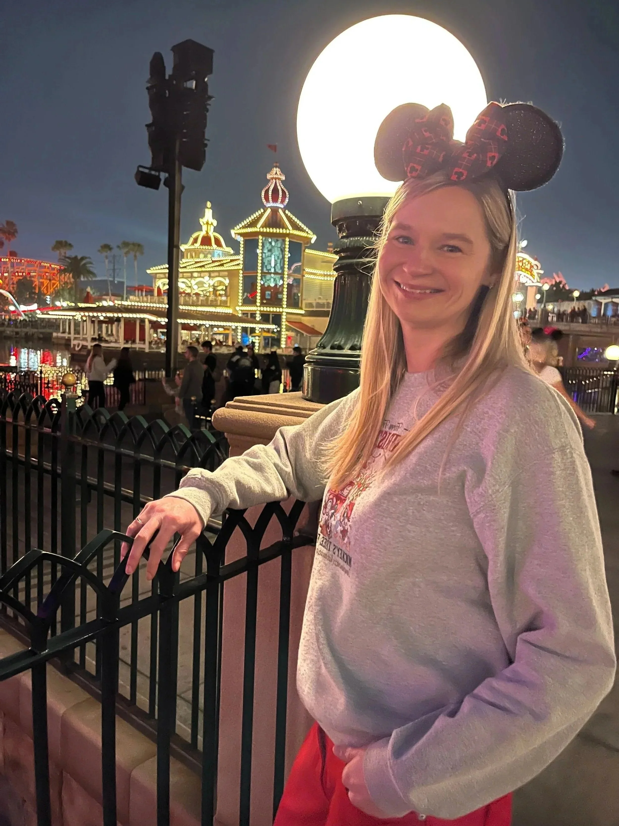 A woman wearing Mickey Mouse ears with red and black patterns smiles at the camera during nighttime at a Disney theme park, with illuminated rides and buildings in the background and a large illuminated lamp behind her.