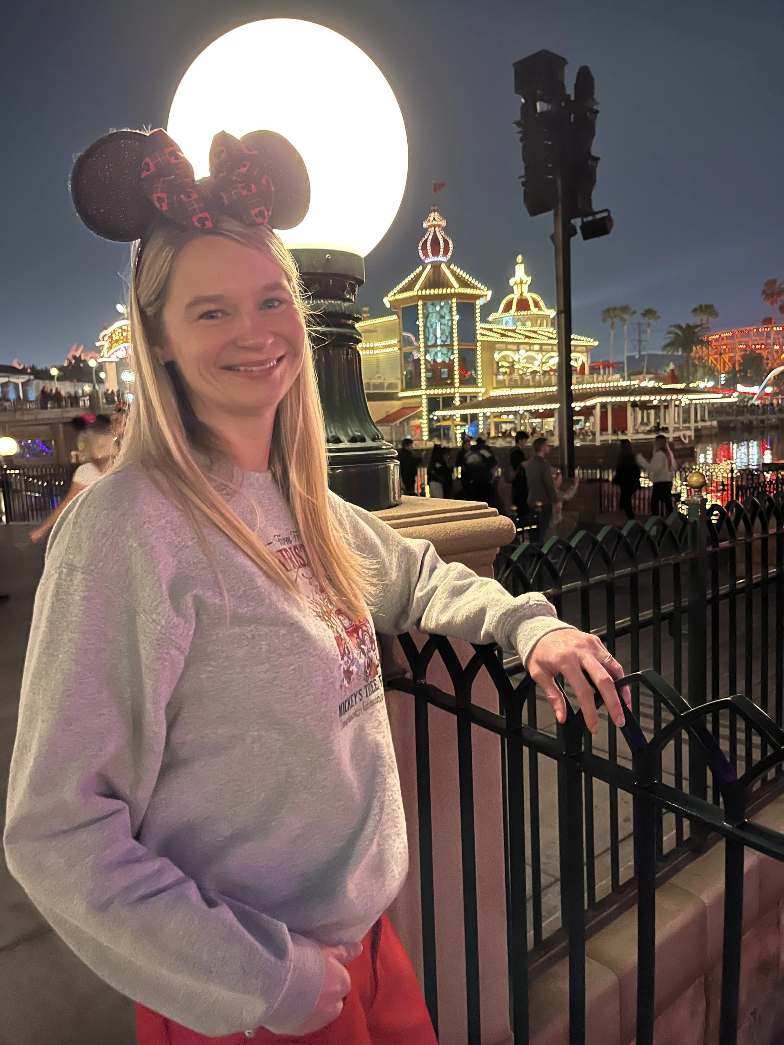 A woman wearing Mickey Mouse ears with red and black patterns, smiling at the camera, standing outdoors at night with a lit-up amusement park in the background, including a large glowing lantern, a castle structure, and a roller coaster, with other visitors visible.