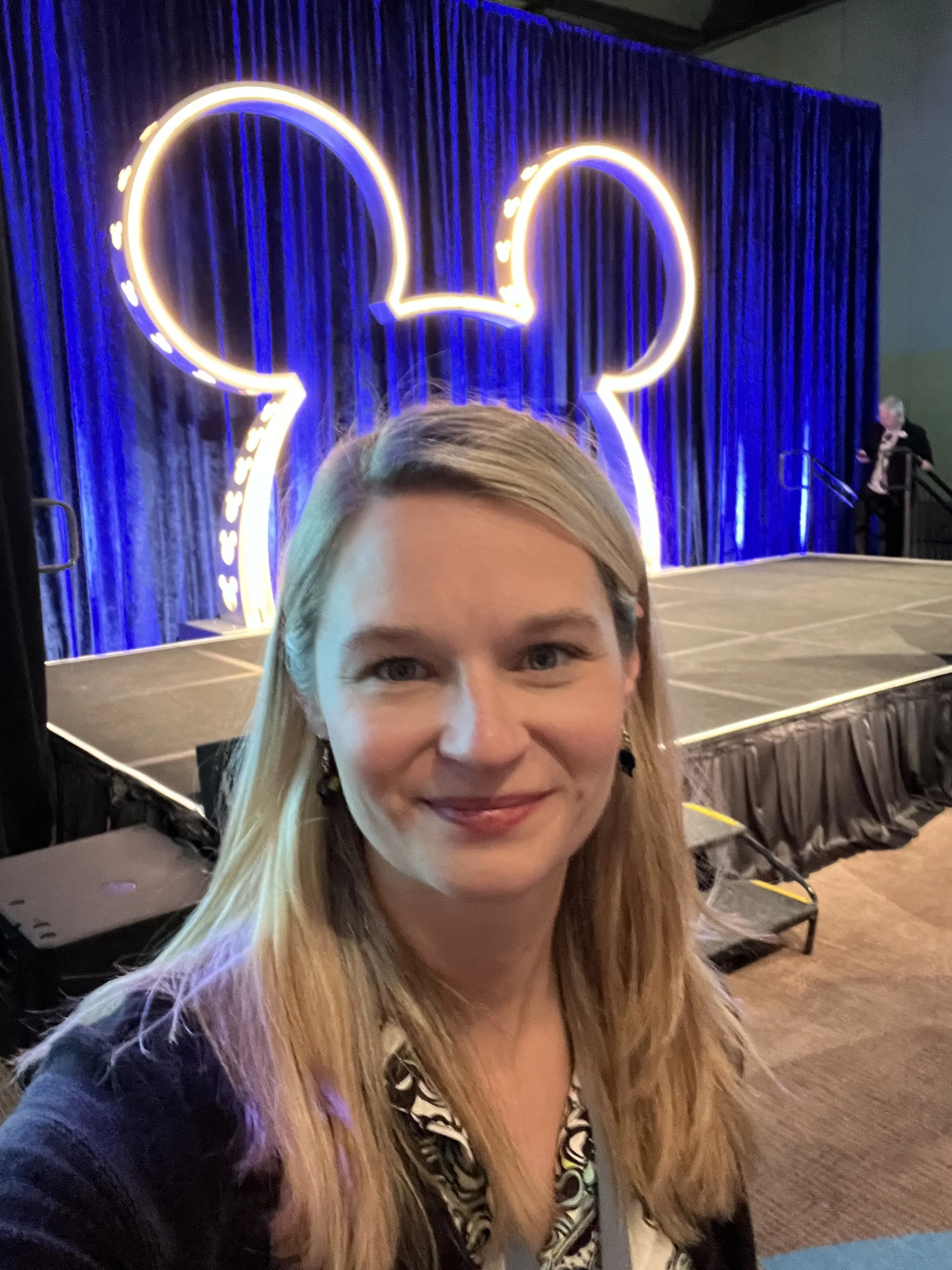 A woman taking a selfie at an event with blue neon lights shaped like Mickey Mouse ears in the background.