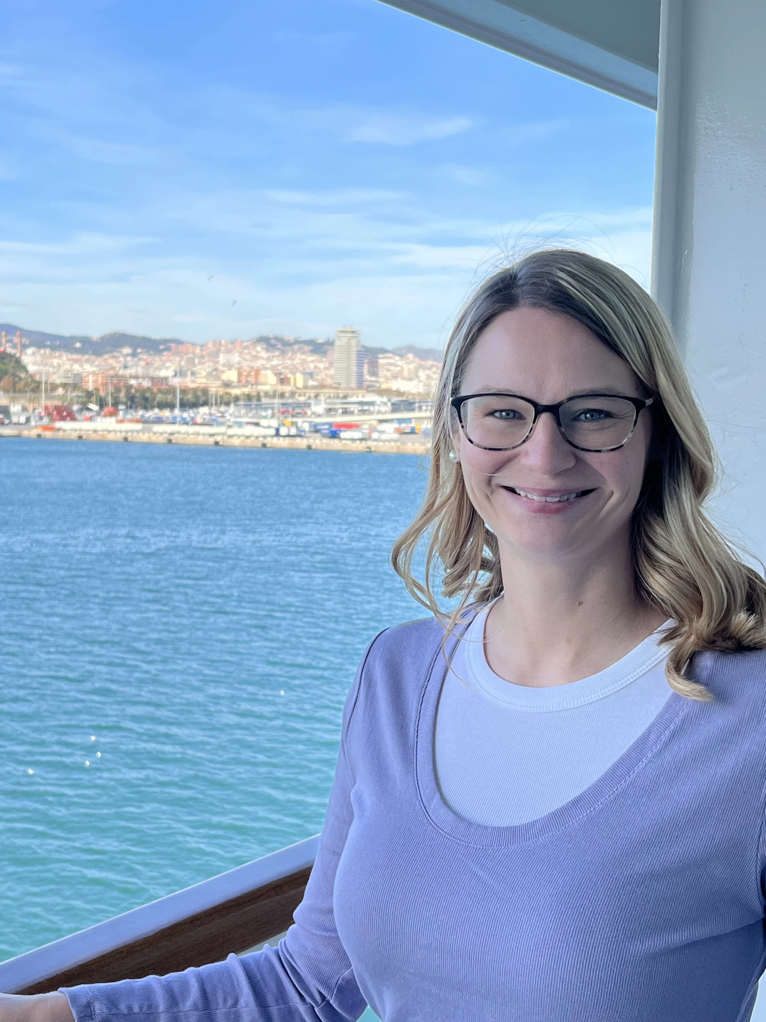 A smiling woman with glasses and blonde hair on a boat with a harbor view, city skyline, and blue sky in the background.