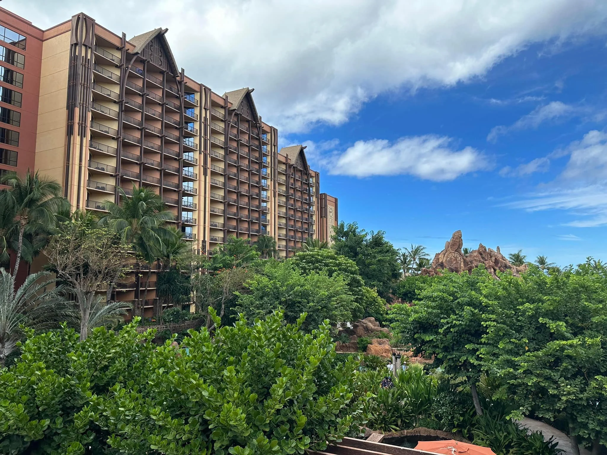 A tall hotel building with multiple balconies surrounded by lush green trees and vegetation, against a blue sky with some clouds.