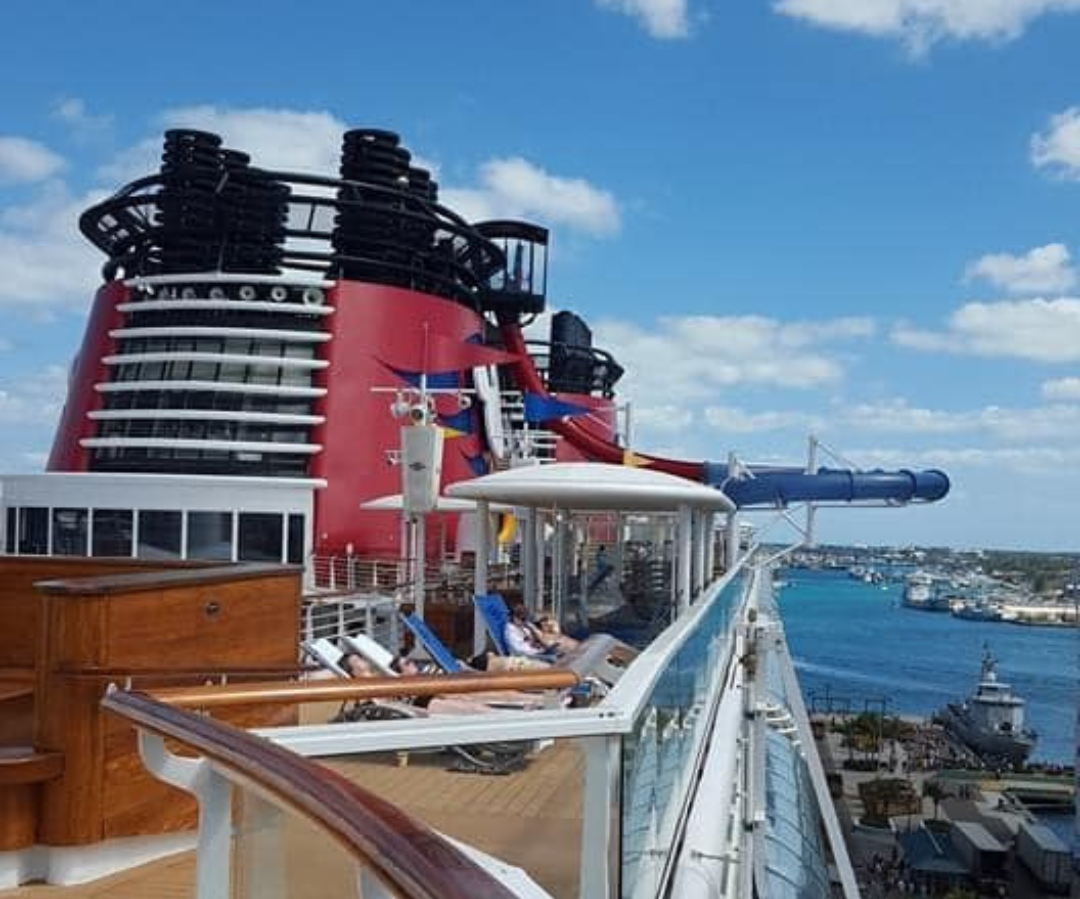 View of the deck area on a cruise ship with lounge chairs, blue skies with clouds, and a cityscape with boats and ships in the water in the background.