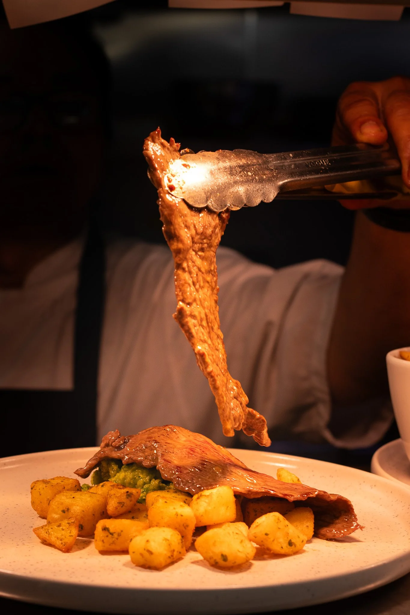 Plate of cooked beef slices with golden roasted potatoes and a green garnish, served on a white round plate. A hand uses tongs to add another slice of beef.