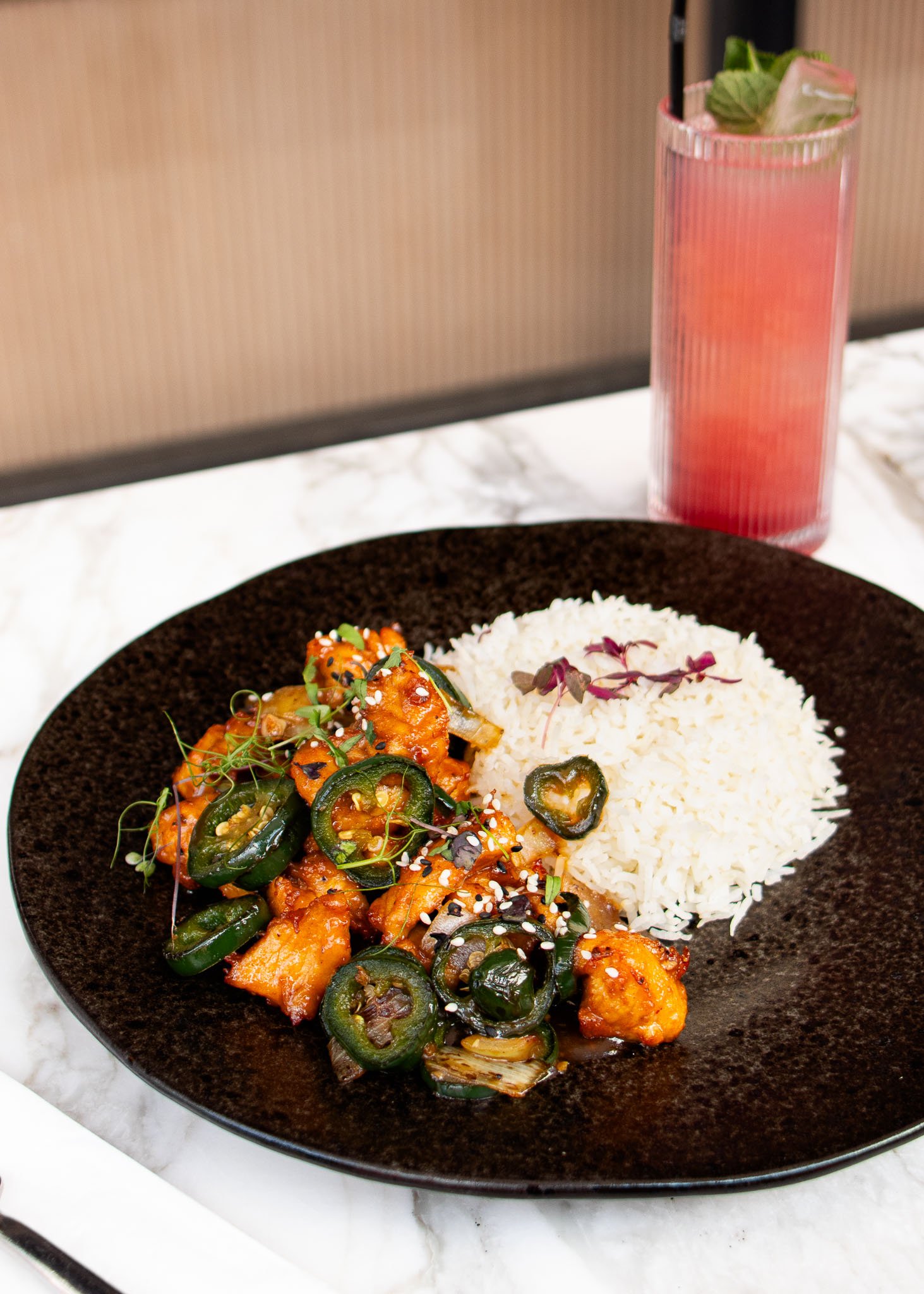 Black plate with rice, spicy chicken, and jalapeños next to a red cocktail with mint garnish on a marble table.