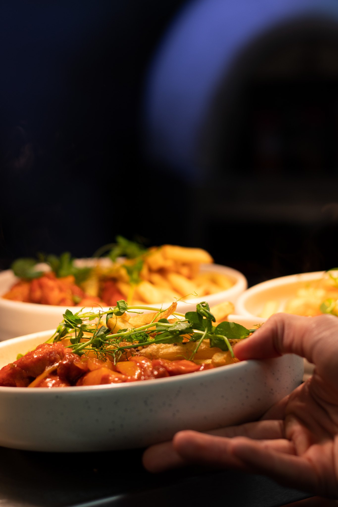 Close-up of a hand holding a white bowl containing food, including fries, leafy greens, and sauced meat or vegetables.