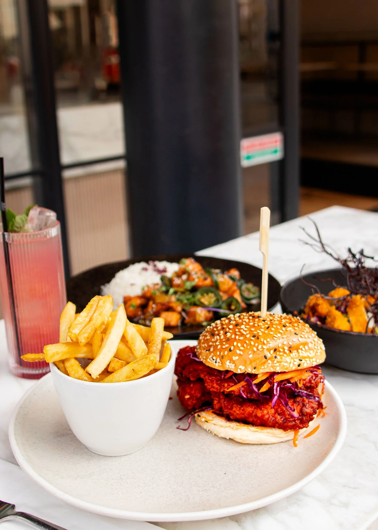 A table with a fried chicken sandwich with slaw on a sesame seed bun, a bowl of French fries, a plate of rice and vegetables, and a pink beverage with ice in a tall glass.