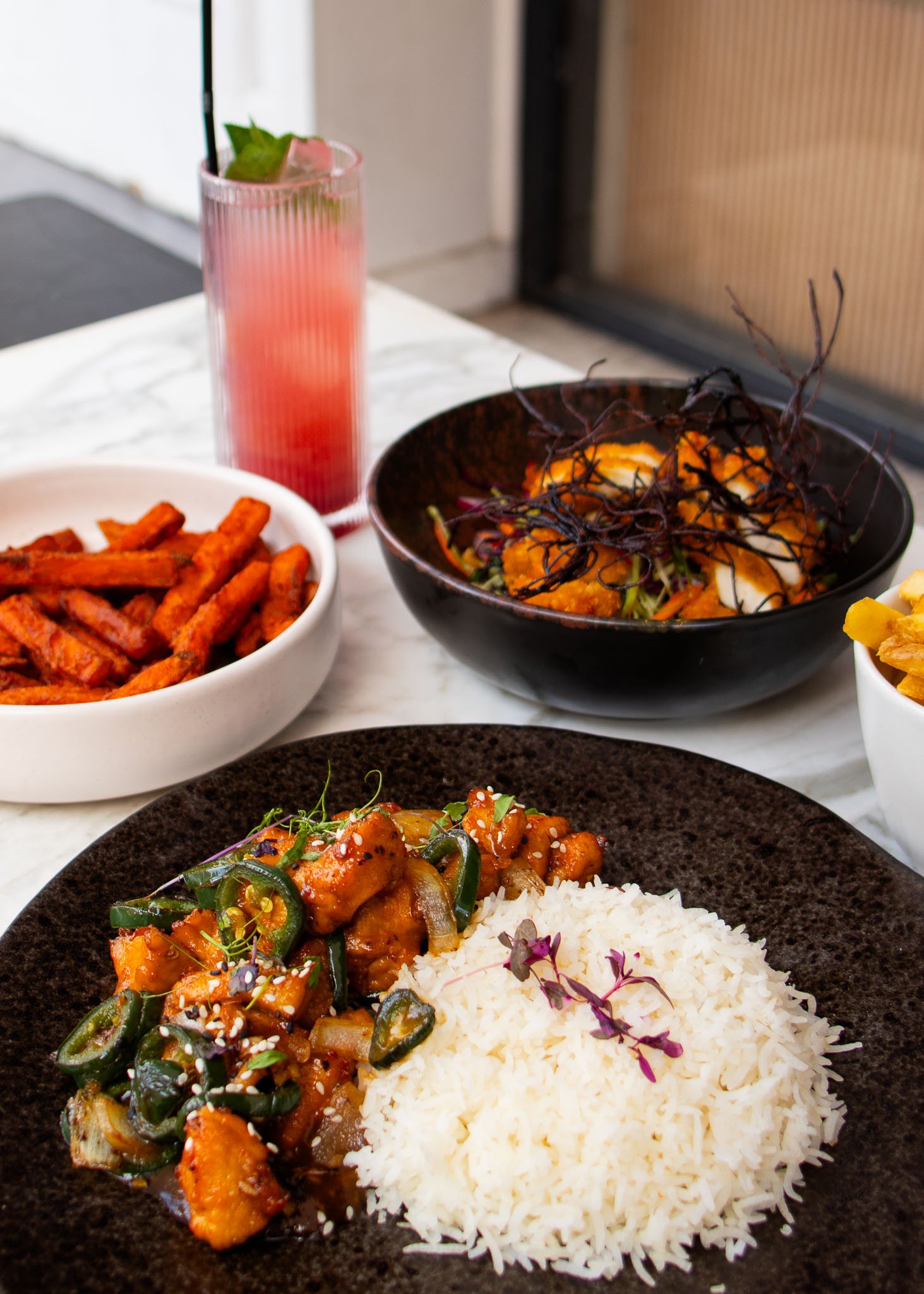 Close-up of a meal with vibrant dishes including a black plate of spicy chicken stir-fry and rice, a bowl of sweet potato fries, a colorful salad, and a pink cocktail with mint in a ribbed glass.