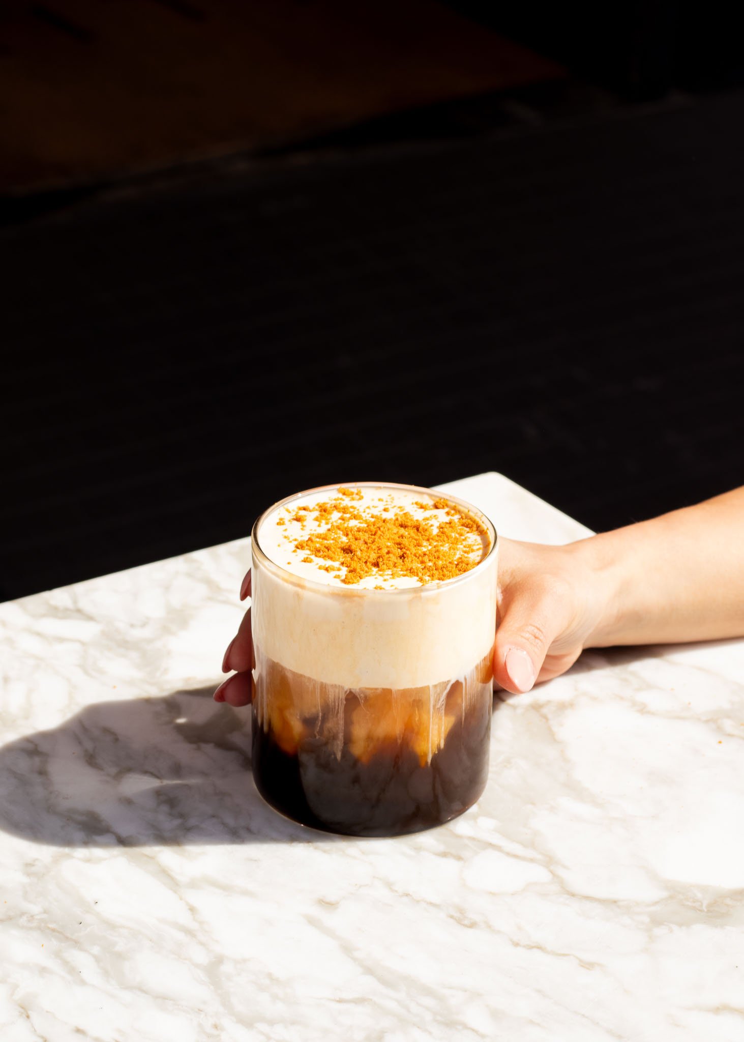 Hand holding a glass of iced coffee with foam and crumbled topping on marble table.