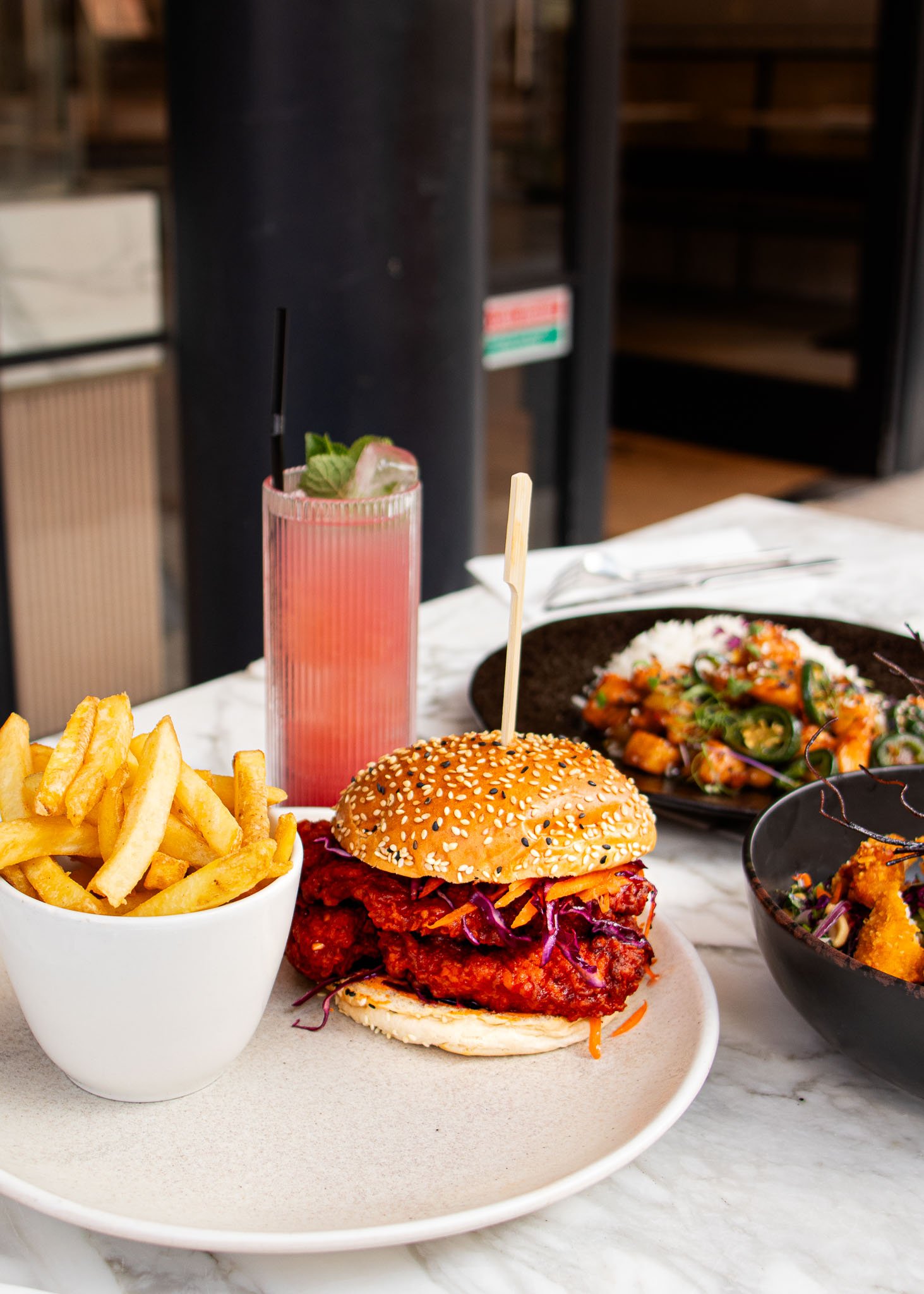 A plate with a sesame seed burger containing fried chicken and coleslaw, accompanied by a bowl of French fries. Next to it is a tall glass of pink lemonade with mint, and a dish of mixed vegetables and rice.