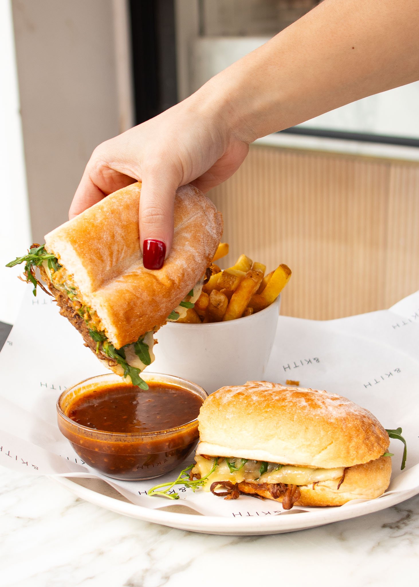 A hand dipping a sandwich into a bowl of sauce next to fries on a table.