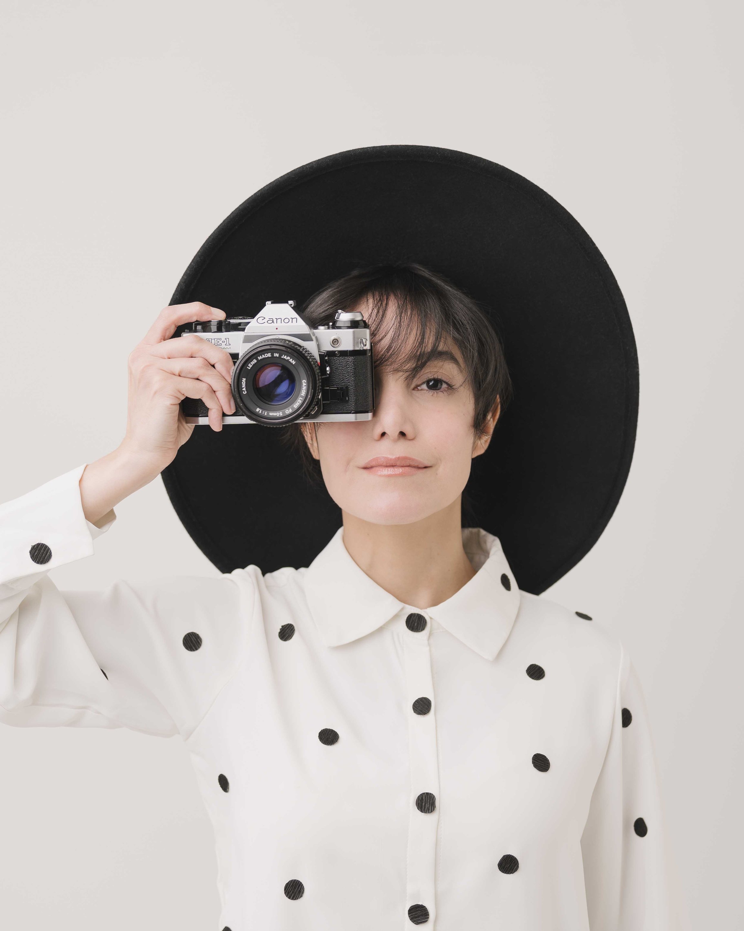 Luisa Del Valle, Charlotte NC children’s photographer, holding a film camera and wearing a black hat in a soft, minimal studio portrait