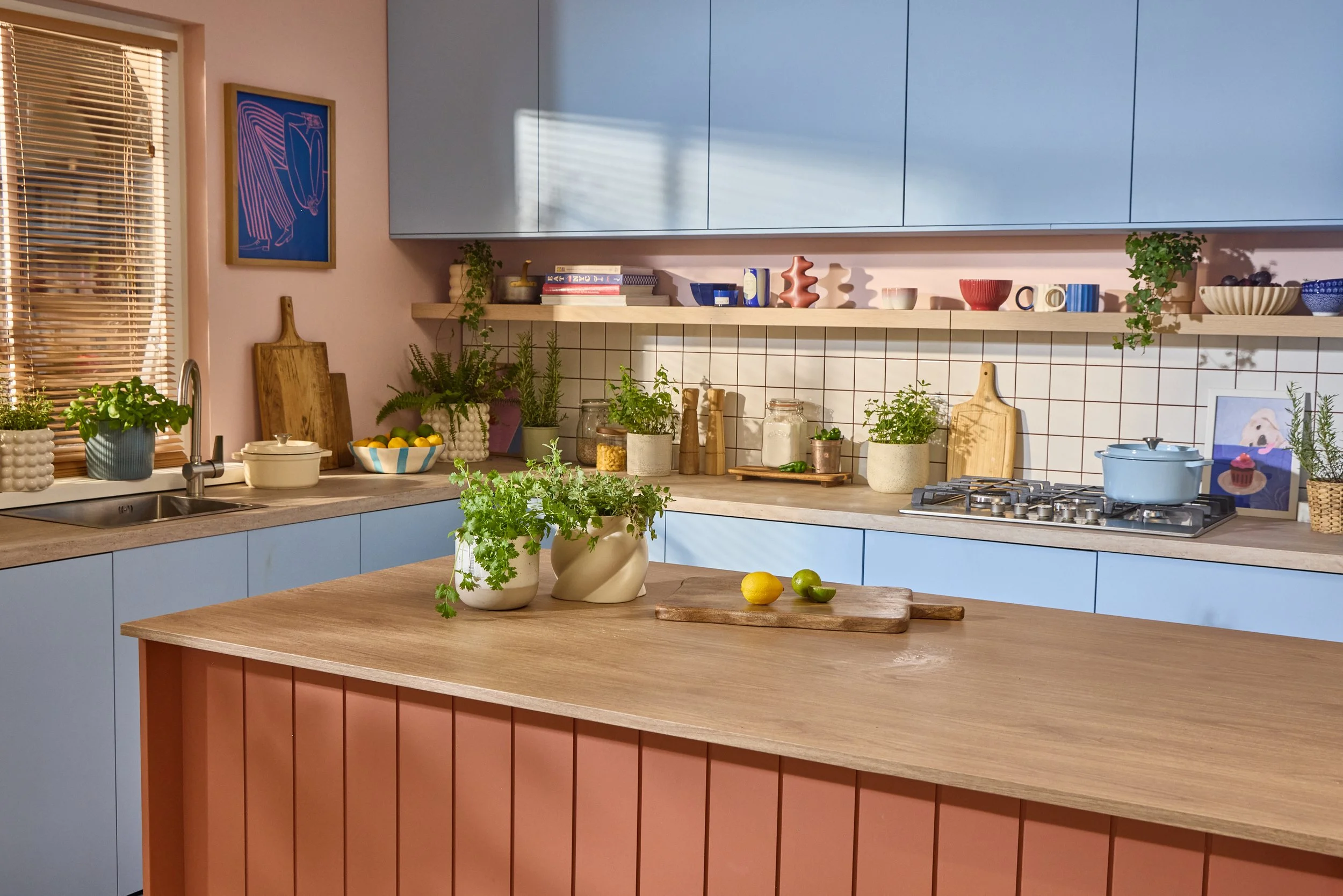 Modern kitchen with pastel blue cabinets, wooden countertops, and a tiled backsplash. Open shelving displays colorful bowls, cups, and decorative items. Countertop has green potted plants, jars, cutting boards, and a bowl of citrus fruits. The foreground features two vases with green plants and a cutting board with lemon and lime.