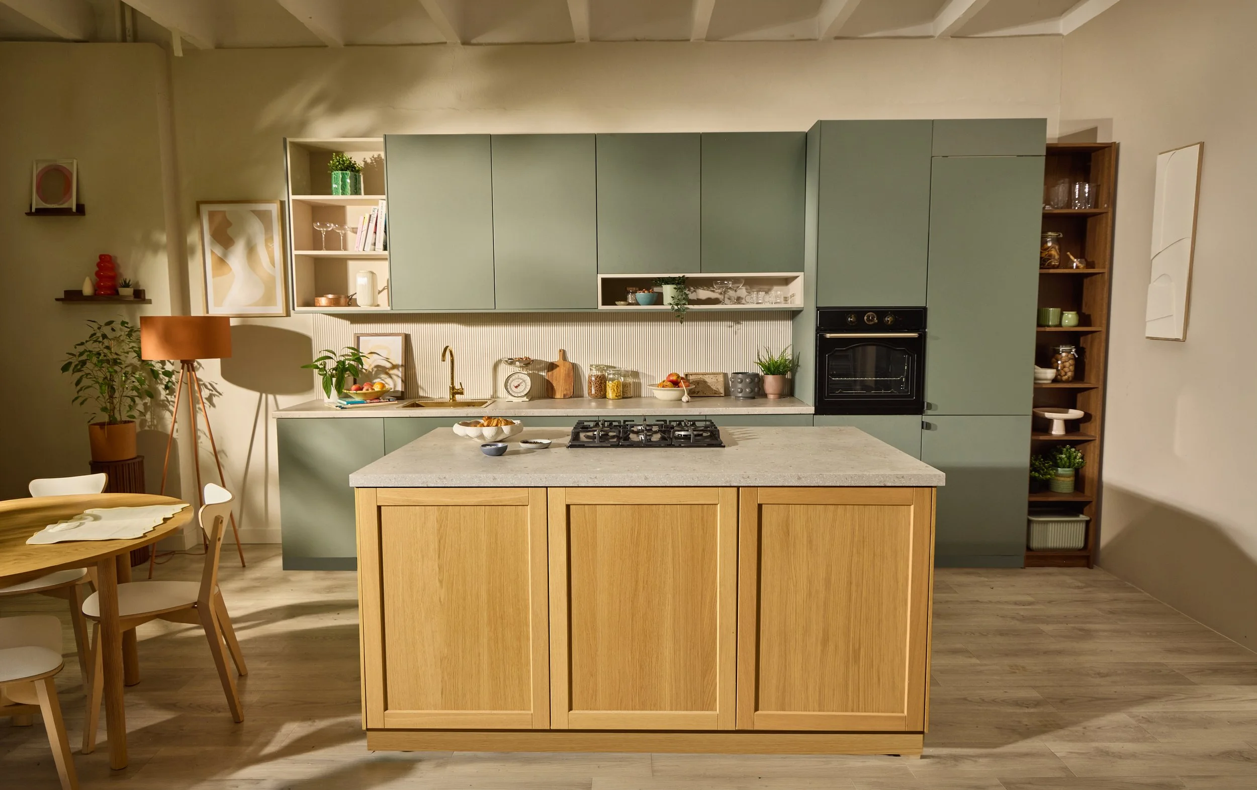 Contemporary kitchen with green cabinets, a wooden island, and open shelving displaying glassware and plants. Adjacent dining area has a round wooden table with chairs, and a tall floor lamp with a reddish-brown shade.