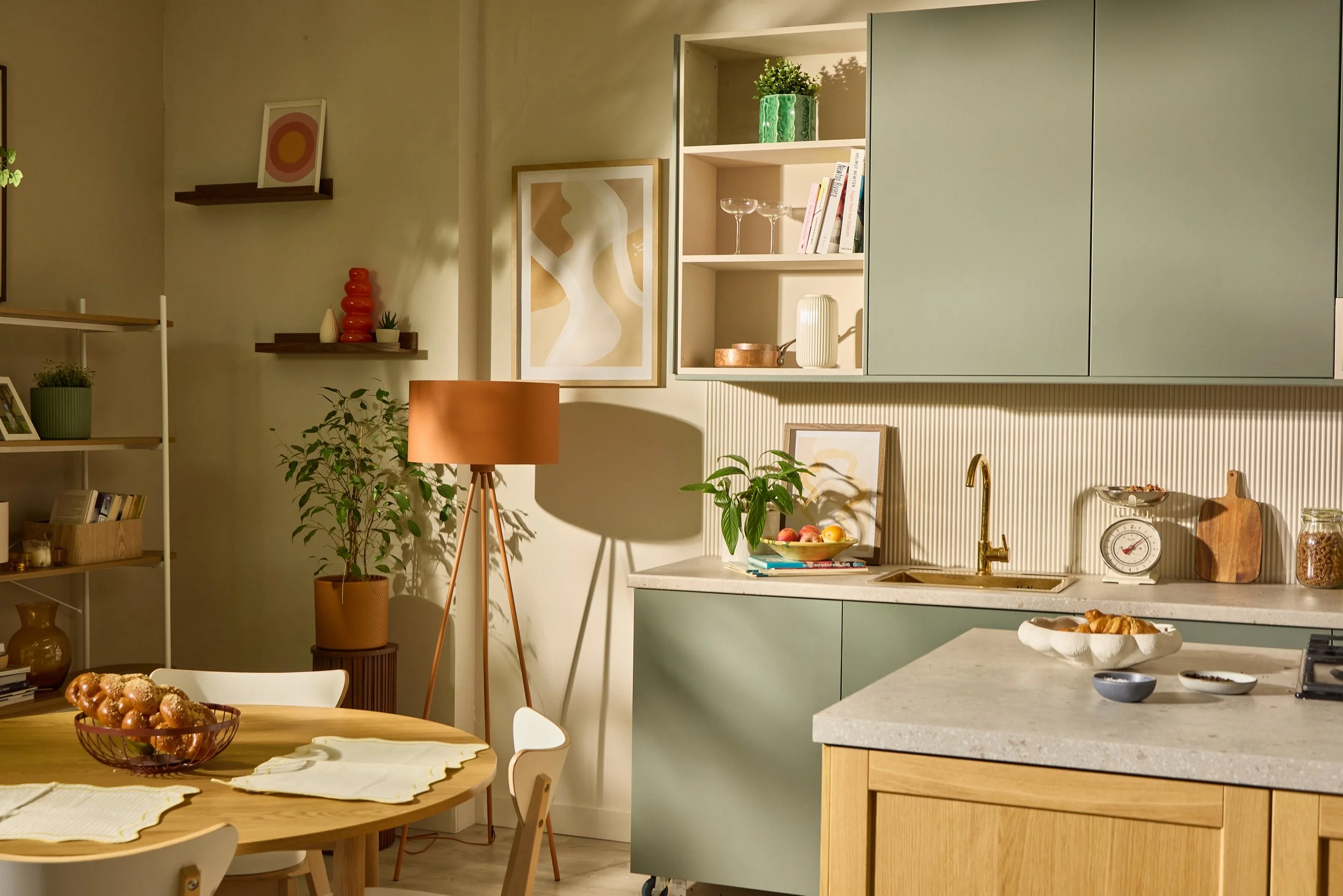 A kitchen with green cabinets, open shelving with glasses and books, a countertop with a sink, a bowl of peaches, and a dining table with a basket of muffins and white chairs.