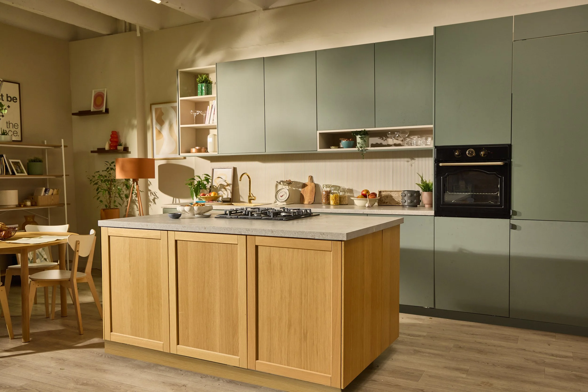 A modern kitchen with a light green upper cabinet, a black oven, a wooden kitchen island, and various decorative items such as plants, bowls, jars, and kitchen tools on the countertop. There is a dining table with white chairs in the foreground and shelves with framed pictures, books, and decorative objects on the left side of the image.