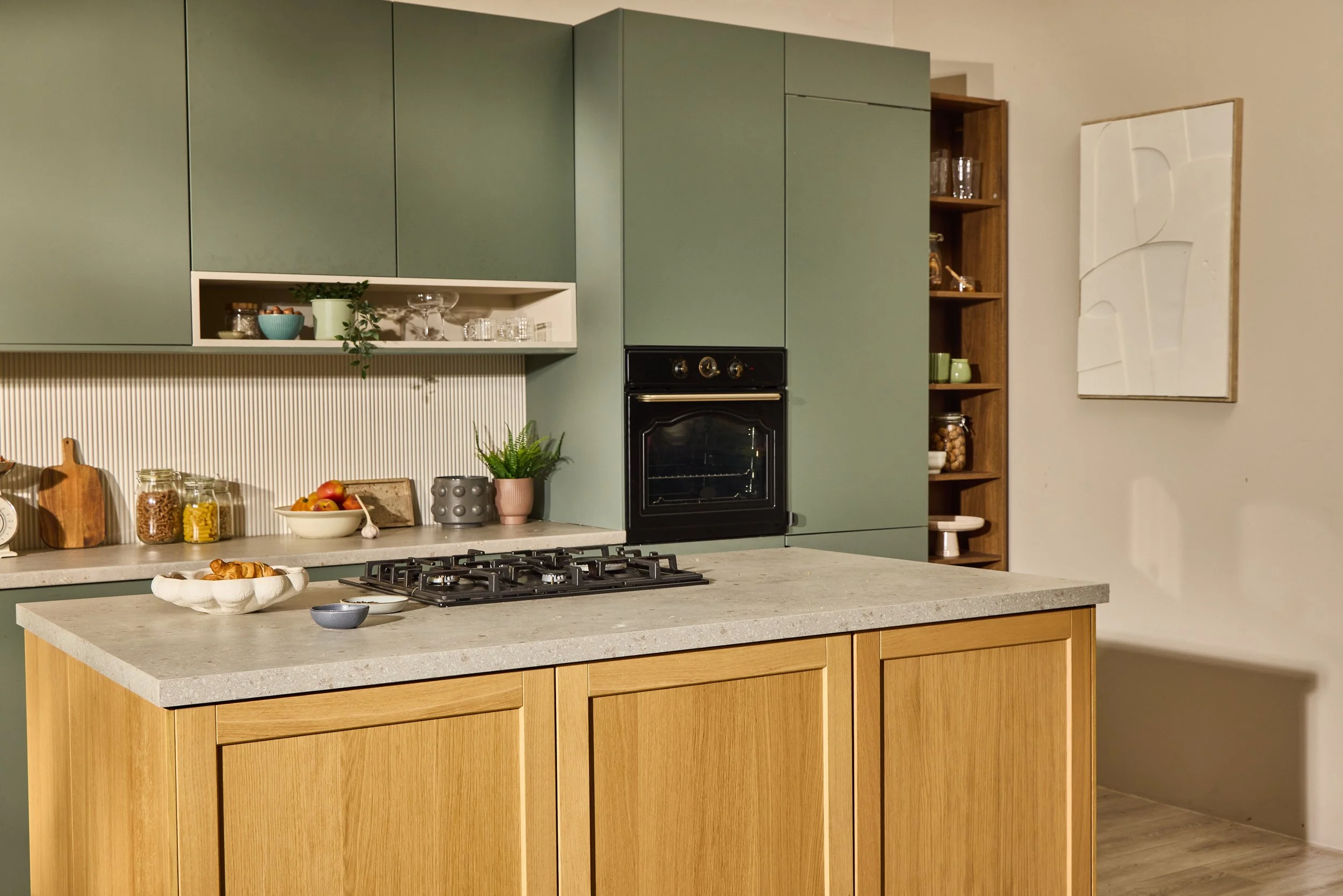 Modern kitchen with wooden island, green cabinets, white countertops, built-in oven, open shelving with glassware, decorative jars, and plants.